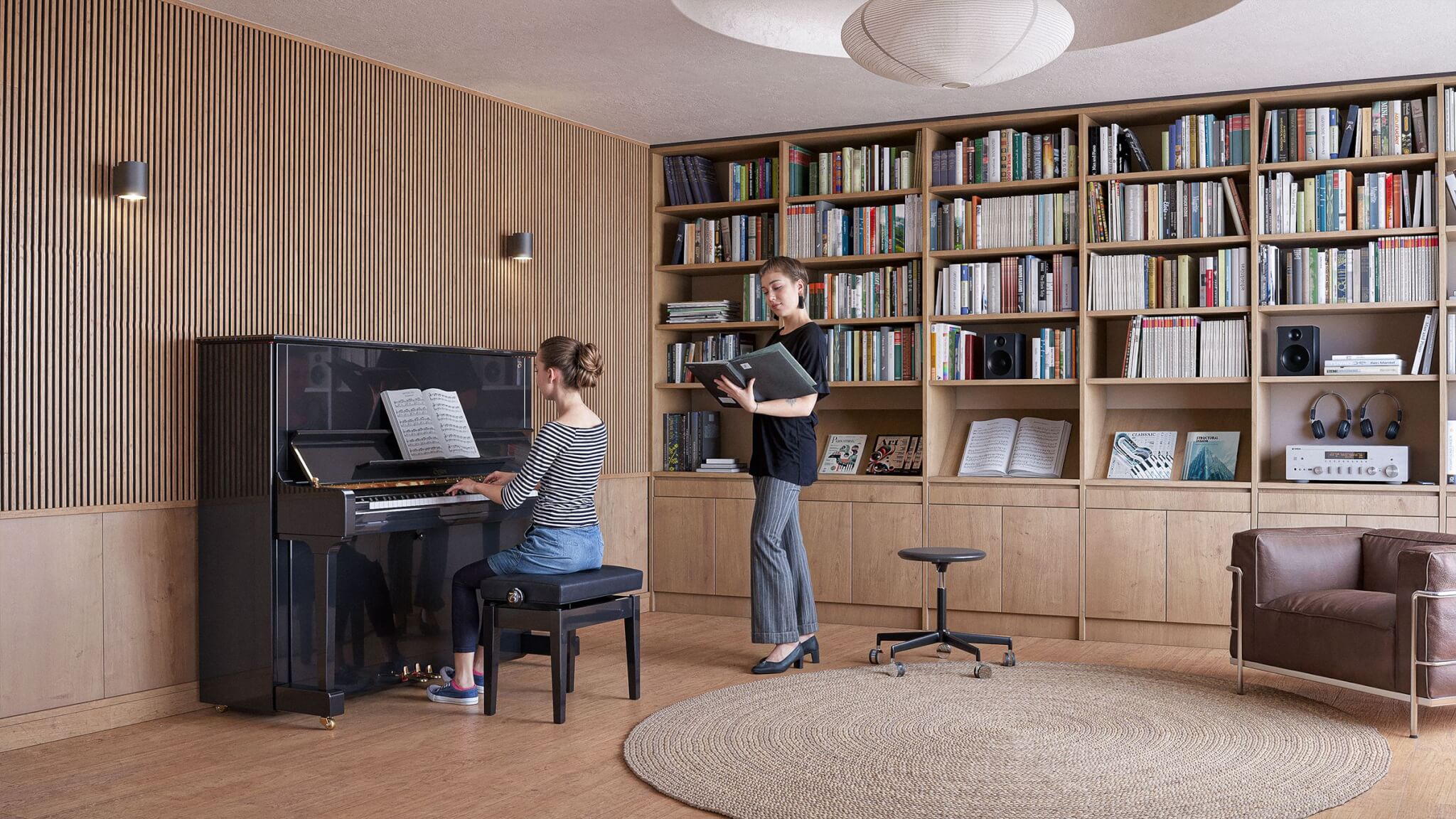 two women inside a room full of books. One of the females is playing the Steinway, while the other lady is looking at a book of notes